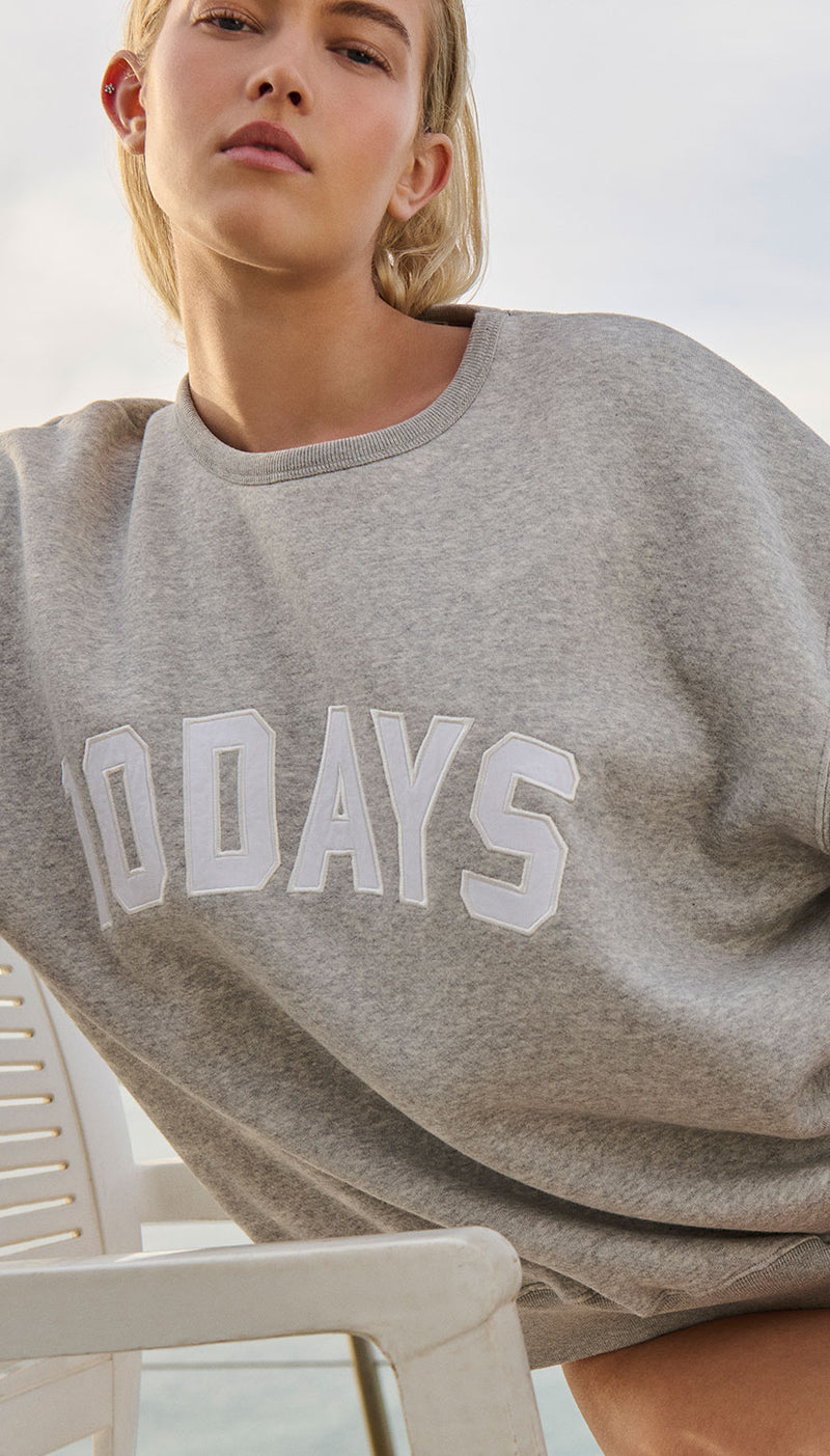 Woman wearing the light grey melee 10DAYS statement sweater. She is sitting on a white chair with a grey blue sky on the background.